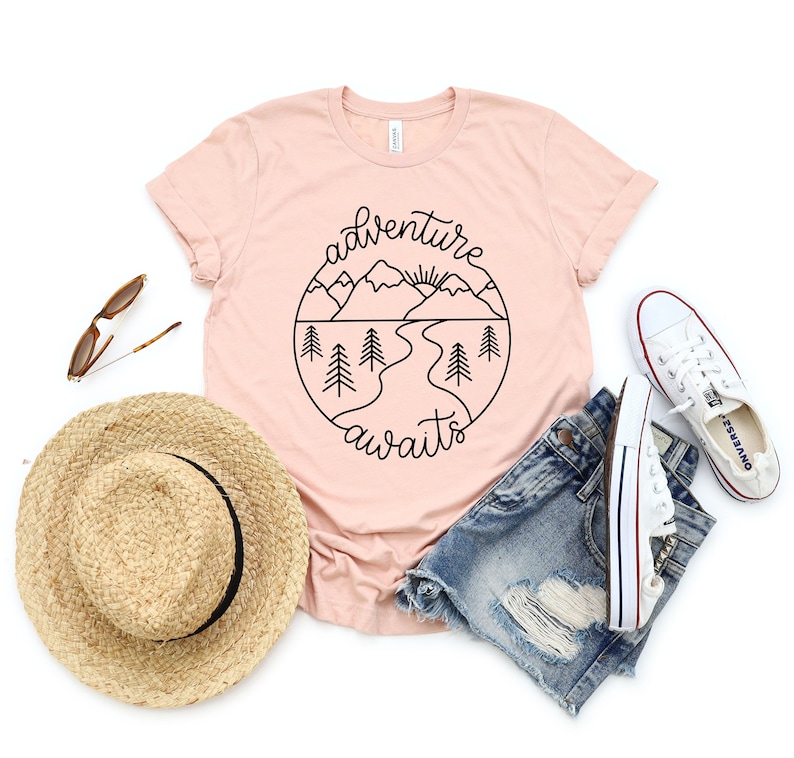 May include: A pink t-shirt with a black graphic of mountains, a river, and a sun. The text "adventure awaits" is written in a cursive font around the graphic.
