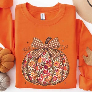 May include: A bright orange crewneck sweatshirt featuring a central graphic of a pumpkin. The pumpkin has a brown and white gingham bow on top and is filled with a colorful floral pattern in shades of orange, red, and brown. Small star accents surround the pumpkin.