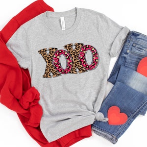 May include: A gray t-shirt with the word "XOXO" printed in pink and brown leopard print. The shirt is folded and displayed with red and blue clothing items.