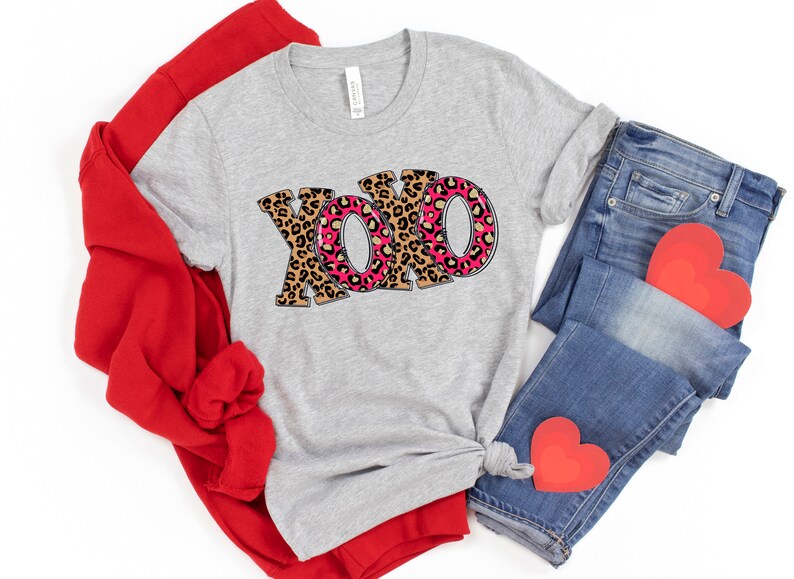 May include: A gray t-shirt with the word "XOXO" printed in pink and brown leopard print. The shirt is folded and displayed with red and blue clothing items.