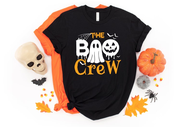 May include: Black t-shirt featuring a white graphic of a ghost and a pumpkin with the text "The Boo Crew". The shirt is surrounded by Halloween decorations, including a skull, bats, candy corn, pumpkins, and autumn leaves.