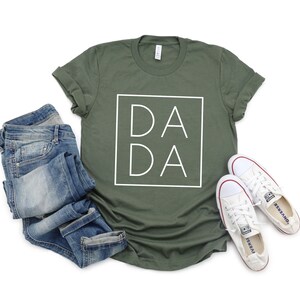 May include: An olive green t-shirt with the word "DADA" in a white square. The shirt is paired with folded blue jeans and white canvas sneakers with red accents.