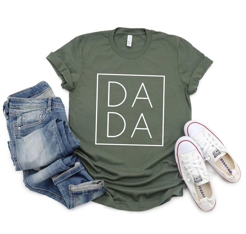 May include: An olive green t-shirt with the word "DADA" in a white square. The shirt is paired with folded blue jeans and white canvas sneakers with red accents.
