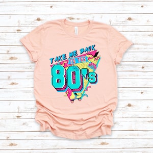 Op de afbeelding: Een roze T-shirt met een grafisch ontwerp met de tekst "Take Me Back To The 80's" in een kleurrijke, retro stijl.
