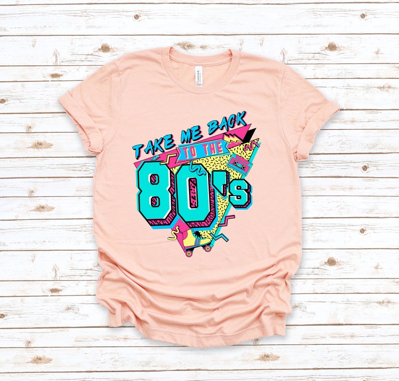 Op de afbeelding: Een roze T-shirt met een grafisch ontwerp met de tekst "Take Me Back To The 80's" in een kleurrijke, retro stijl.