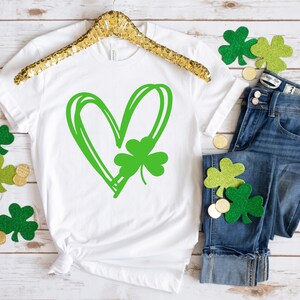 May include: White t-shirt with a green shamrock inside a green heart design. The shirt is laid flat on a white wooden surface with green shamrock decorations and gold coins.