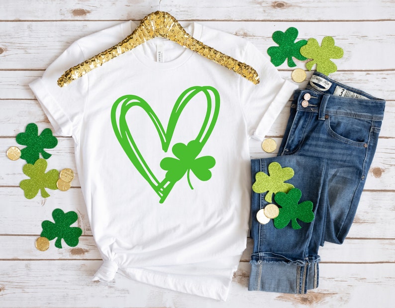 May include: White t-shirt with a green shamrock inside a green heart design. The shirt is laid flat on a white wooden surface with green shamrock decorations and gold coins.