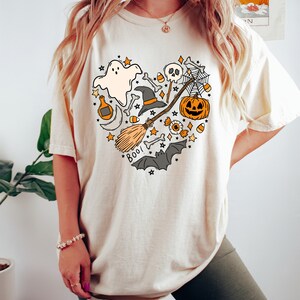 Może przedstawiać: Kremowa koszulka z nadrukiem w kształcie serca, przedstawiającym r&oacute;żne symbole Halloween, w tym ducha, kapelusz czarownicy, miotłę, dynię, czaszkę, cukrowe kukurydzę i nietoperze. Na koszulce widnieje napis "Boo!"