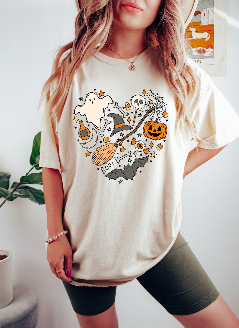 Może przedstawiać: Kremowa koszulka z nadrukiem w kształcie serca, przedstawiającym r&oacute;żne symbole Halloween, w tym ducha, kapelusz czarownicy, miotłę, dynię, czaszkę, cukrowe kukurydzę i nietoperze. Na koszulce widnieje napis "Boo!"