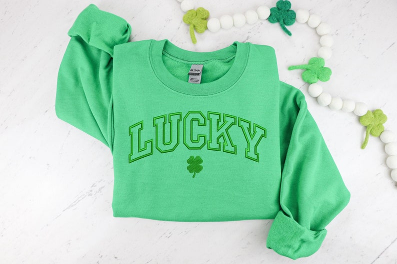 K&ouml;nnte beinhalten: Ein gr&uuml;ner Pullover mit dem Wort "LUCKY" in wei&szlig;er Umrandung und einem gr&uuml;nen Kleeblatt darunter.