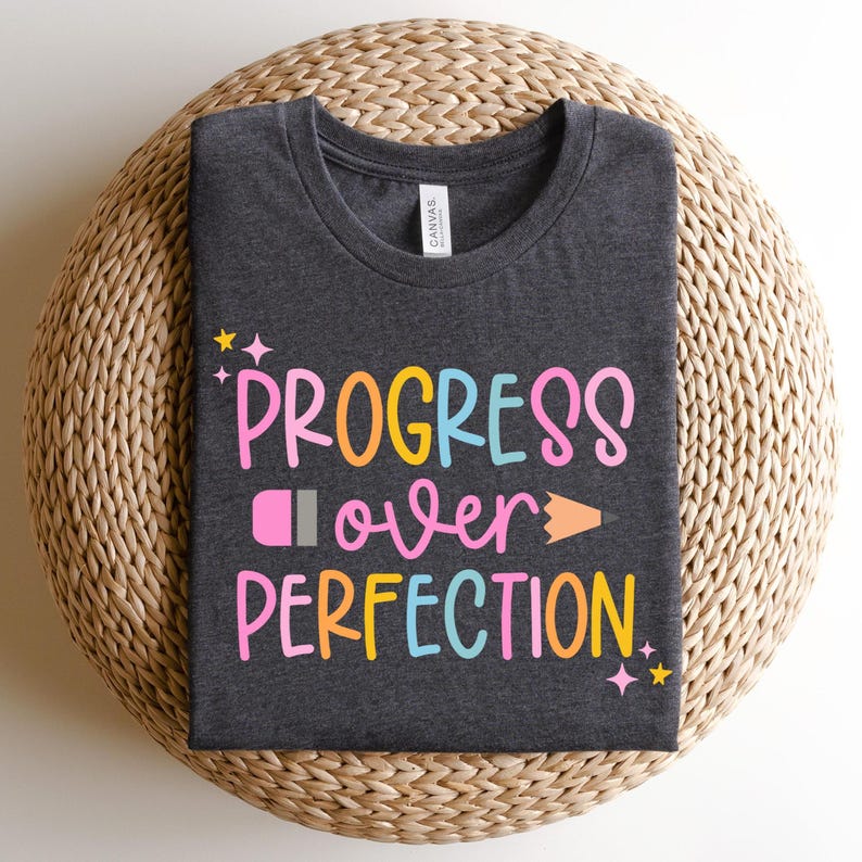 May include: Dark gray t-shirt with the words "PROGRESS over PERFECTION" in colorful, playful lettering. A pencil graphic is incorporated into the design. The shirt is folded and resting on a woven, circular surface.