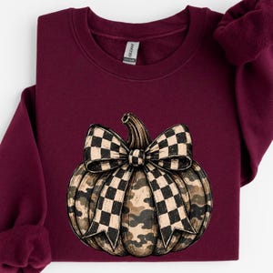 May include: A maroon long-sleeve sweatshirt featuring a central graphic of a pumpkin with a camouflage pattern and a large black and white checkered bow. The garment has a crew neckline and a Gildan brand label visible at the inner collar.