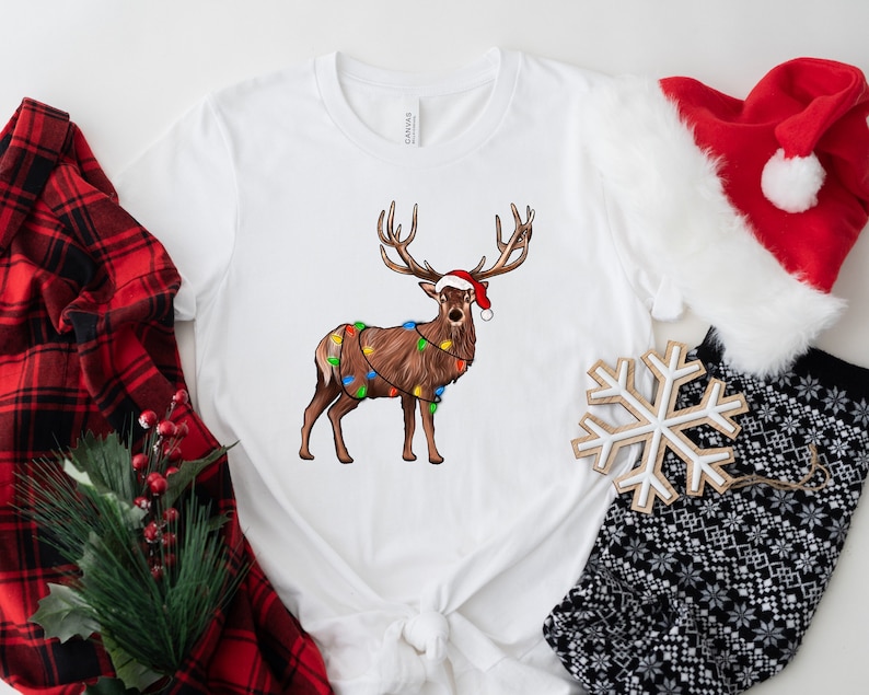 Puede incluir: Camiseta blanca con un reno marr&oacute;n que lleva un gorro de Pap&aacute; Noel rojo y est&aacute; envuelto en luces de Navidad de colores.