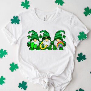 Peut inclure: T-shirt blanc avec un motif de trois lutins portant des chapeaux verts et tenant un fer &agrave; cheval, un arc-en-ciel et un tr&egrave;fle. Les lutins portent des chemises ray&eacute;es vertes et blanches et ont des barbes blanches.