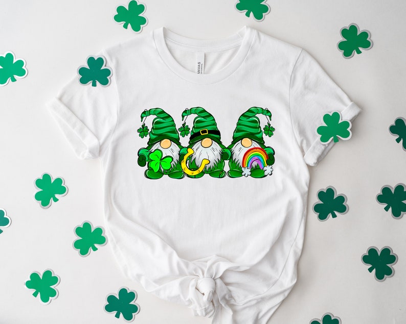 Peut inclure: T-shirt blanc avec un motif de trois lutins portant des chapeaux verts et tenant un fer &agrave; cheval, un arc-en-ciel et un tr&egrave;fle. Les lutins portent des chemises ray&eacute;es vertes et blanches et ont des barbes blanches.