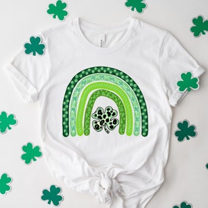 May include: A white t-shirt with a green shamrock rainbow design. The rainbow is made up of five green stripes with different patterns, including shamrocks, dots, and swirls. A large shamrock with a black and white leopard print is in the center of the rainbow.