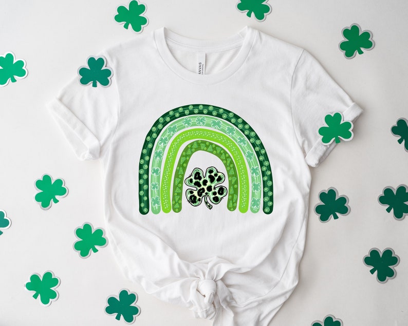 May include: A white t-shirt with a green shamrock rainbow design. The rainbow is made up of five green stripes with different patterns, including shamrocks, dots, and swirls. A large shamrock with a black and white leopard print is in the center of the rainbow.
