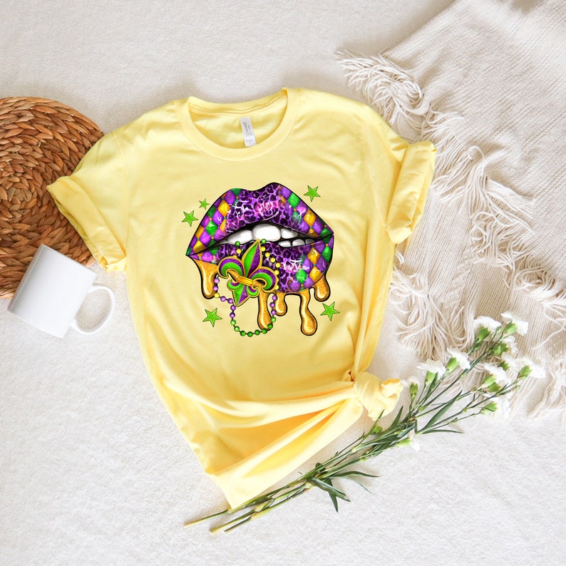 May include: Yellow t-shirt featuring a purple and green Mardi Gras lips design. The lips are dripping with gold paint and have a fleur-de-lis charm hanging from them.