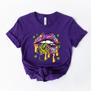 May include: Purple t-shirt featuring a graphic of a dripping, open mouth with a Mardi Gras design. The mouth is decorated with purple, green, and gold colours, and a fleur-de-lis.