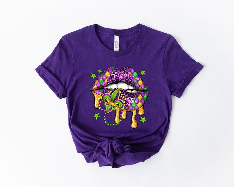 May include: Purple t-shirt featuring a graphic of a dripping, open mouth with a Mardi Gras design. The mouth is decorated with purple, green, and gold colours, and a fleur-de-lis.