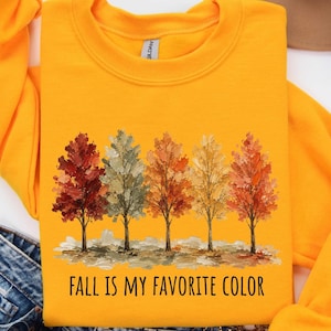 Pode incluir: Uma camisola de gola redonda laranja brilhante com um gráfico de cinco árvores com folhas texturizadas e pictóricas em tons de vermelho, laranja, amarelo e verde suave. Abaixo das árvores, o texto "FALL IS MY FAVORITE COLOR" em preto.