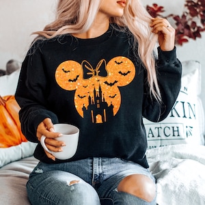 May include: Black sweatshirt featuring a Halloween-themed design. The design includes a Minnie Mouse silhouette with a bow, bats, and a castle in orange glitter. A white mug is held in the hand.