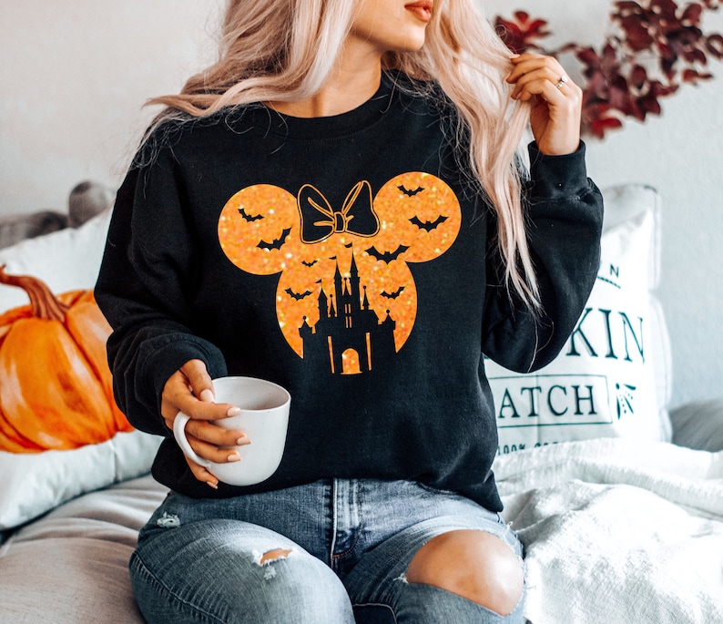 May include: Black sweatshirt featuring a Halloween-themed design. The design includes a Minnie Mouse silhouette with a bow, bats, and a castle in orange glitter. A white mug is held in the hand.