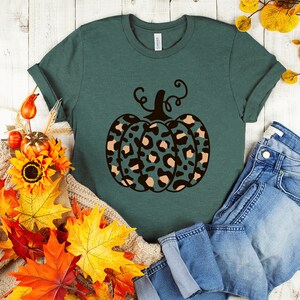 May include: A teal green t-shirt with a black and brown leopard print pumpkin design. The pumpkin has a stem and leaves. The shirt is laying on a white wooden surface with fall leaves and a pair of blue jeans.