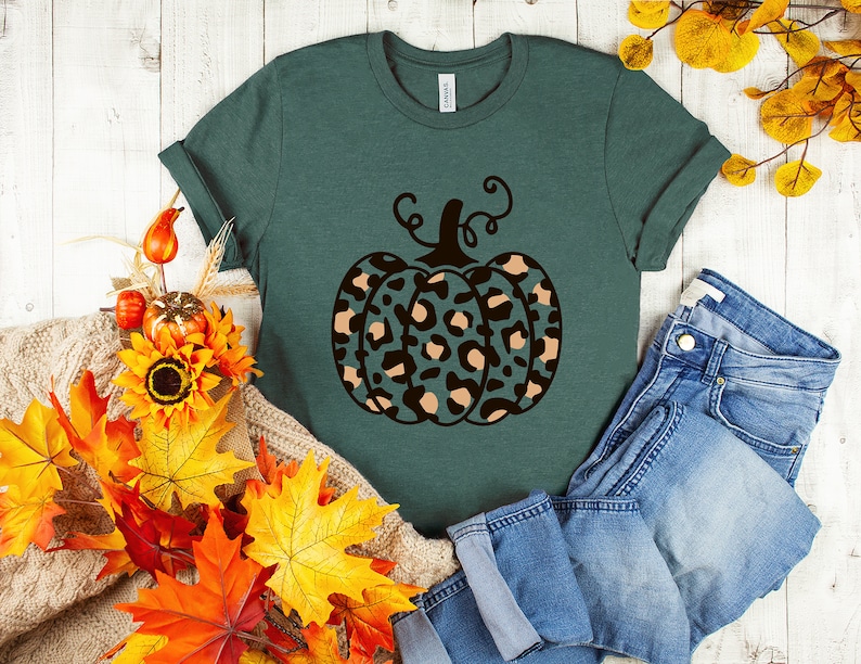 May include: A teal green t-shirt with a black and brown leopard print pumpkin design. The pumpkin has a stem and leaves. The shirt is laying on a white wooden surface with fall leaves and a pair of blue jeans.