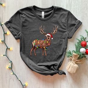 Puede incluir: Una camiseta gris oscuro con un reno de dibujos animados que lleva un gorro de Pap&aacute; Noel y est&aacute; envuelto en luces de Navidad.