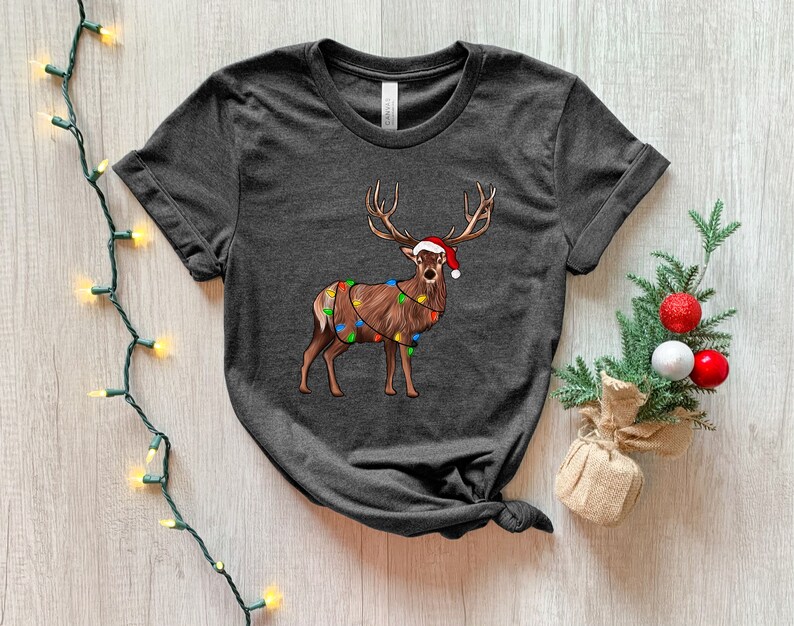 Puede incluir: Una camiseta gris oscuro con un reno de dibujos animados que lleva un gorro de Pap&aacute; Noel y est&aacute; envuelto en luces de Navidad.