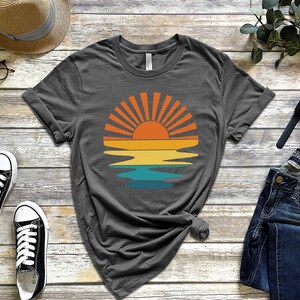 May include: A dark gray t-shirt with a retro-style sunset graphic featuring a sun with rays and a reflection in the water. The graphic is in shades of orange, yellow, blue, and green.