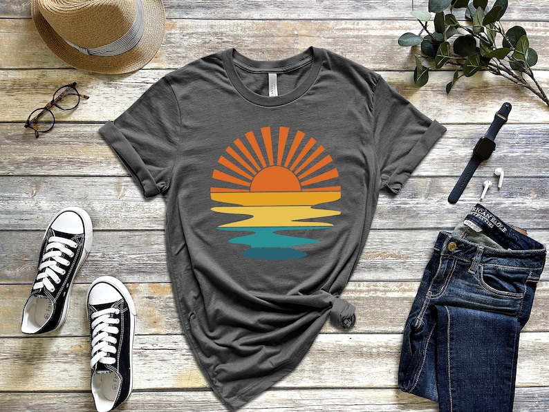 May include: A dark gray t-shirt with a retro-style sunset graphic featuring a sun with rays and a reflection in the water. The graphic is in shades of orange, yellow, blue, and green.