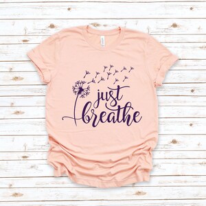 Op de afbeelding: Een perzikkleurig t-shirt met een paarse paardenbloem afbeelding en de tekst "just breathe" in een cursief schrift.