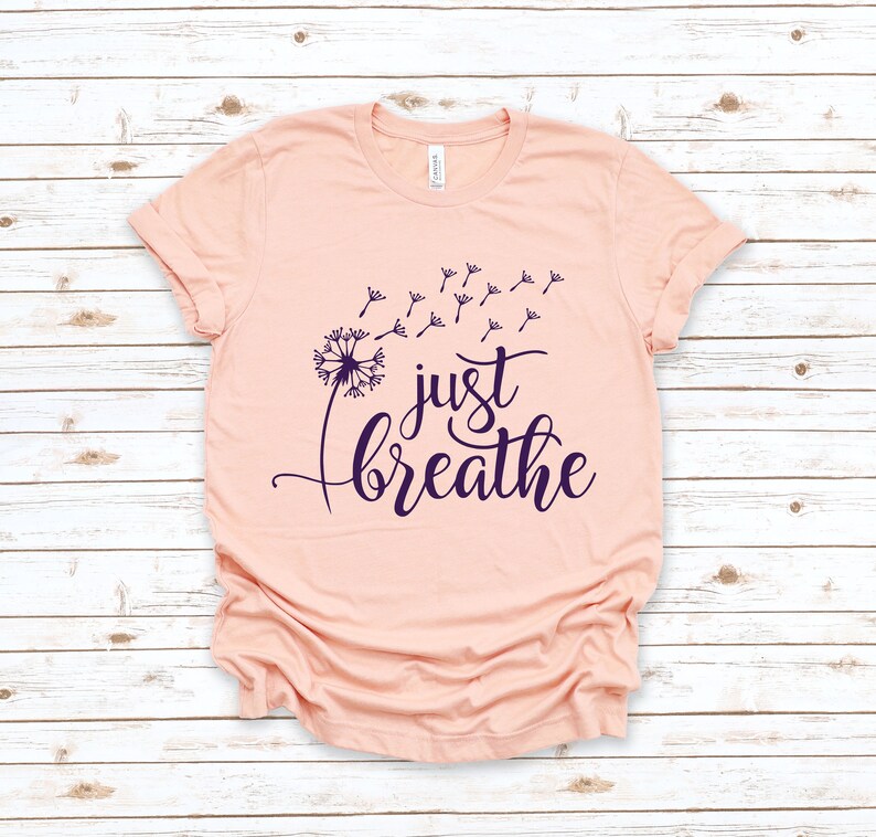 Op de afbeelding: Een perzikkleurig t-shirt met een paarse paardenbloem afbeelding en de tekst "just breathe" in een cursief schrift.