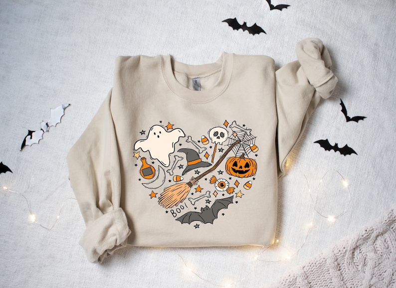 Może przedstawiać: Beżowa bluza z sercowatym wzorem przedstawiającym r&oacute;żne symbole Halloween, w tym ducha, kapelusz czarownicy, miotłę, dynię, czaszkę i napis "BOO!"