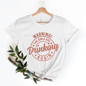May include: White t-shirt with the text "WARNING! THE GIRLS ARE DRINKING AGAIN" in a circular design. The text is in a reddish-brown color. The shirt is hanging on a wooden hanger.