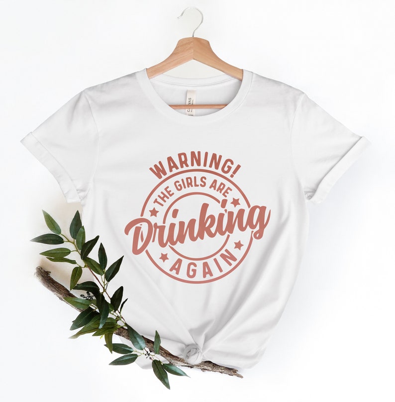 May include: White t-shirt with the text "WARNING! THE GIRLS ARE DRINKING AGAIN" in a circular design. The text is in a reddish-brown color. The shirt is hanging on a wooden hanger.