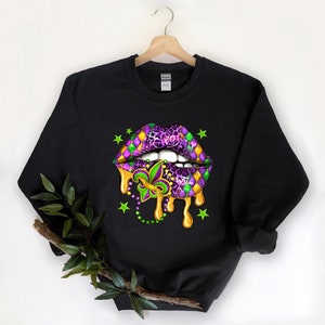 May include: Black sweatshirt featuring a purple and green Mardi Gras lips design. The lips are dripping gold paint and have a fleur-de-lis in the centre. There are green stars and beads around the lips.