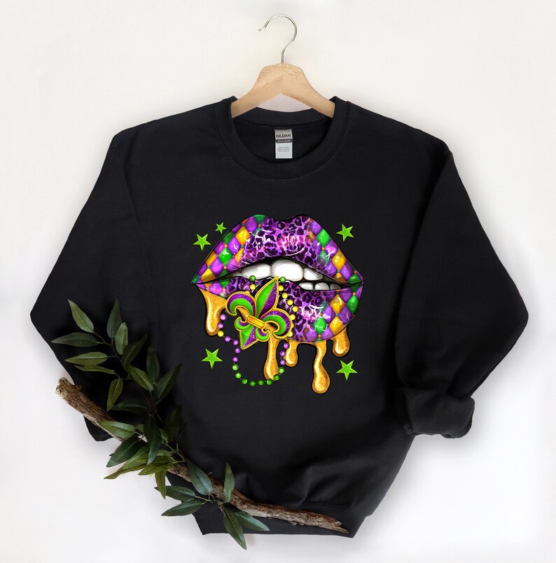 May include: Black sweatshirt featuring a purple and green Mardi Gras lips design. The lips are dripping gold paint and have a fleur-de-lis in the centre. There are green stars and beads around the lips.
