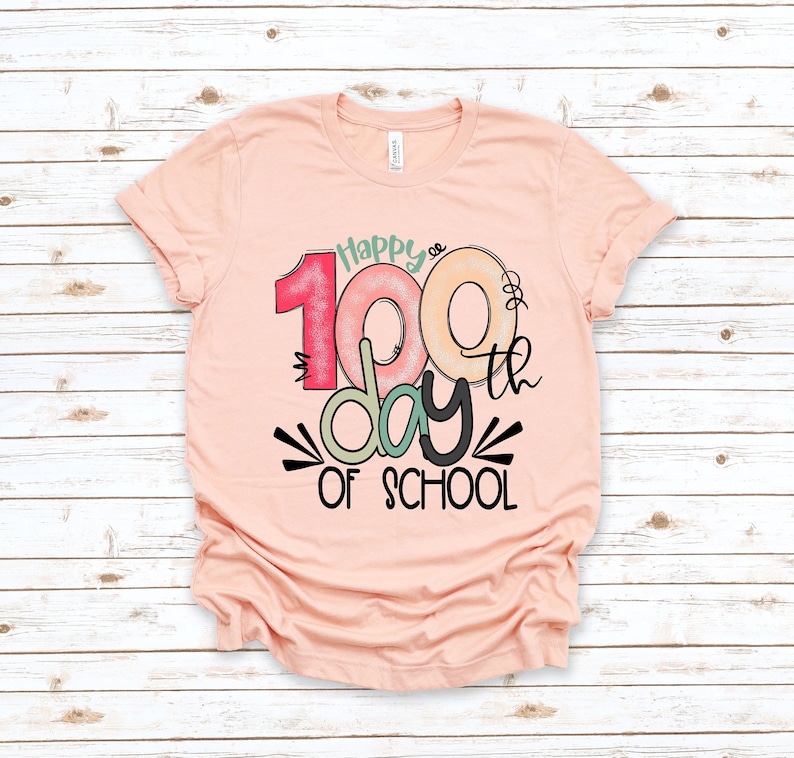 May include: A peach-colored t-shirt with a graphic design that says "Happy 100th day of school". The number 100 is in a colorful donut shape.