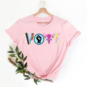 May include: Pink t-shirt with the word "VOTE" spelled out in colorful letters and symbols. The "V" is made of books, the "O" is a fist in a blue circle, the "T" is a pink uterus with flowers, and the "E" is a rainbow.