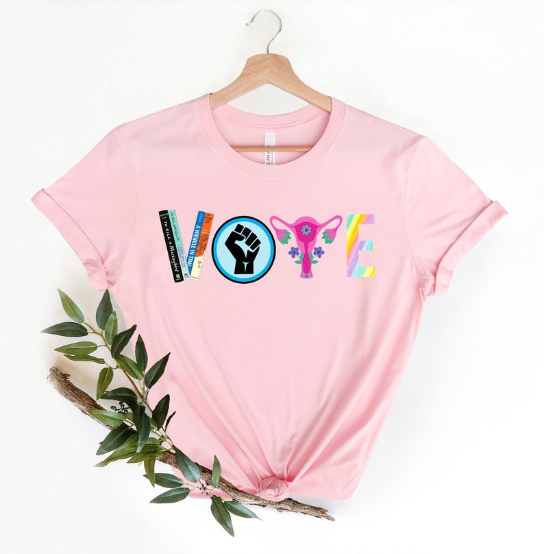 May include: Pink t-shirt with the word "VOTE" spelled out in colorful letters and symbols. The "V" is made of books, the "O" is a fist in a blue circle, the "T" is a pink uterus with flowers, and the "E" is a rainbow.