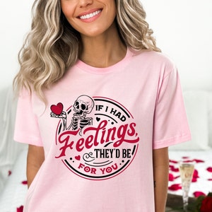 May include: Pink t-shirt featuring a black and white skeleton holding a red heart. The shirt has the text "If I had feelings they'd be for you" in a black circle with a red heart.