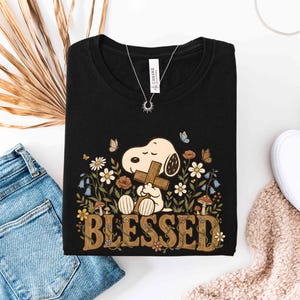 Puede incluir: Camiseta negra con Snoopy sosteniendo una cruz, rodeado de flores y mariposas. La palabra "BLESSED" se muestra en una fuente decorativa. Se ve un collar plateado con un colgante de sol.