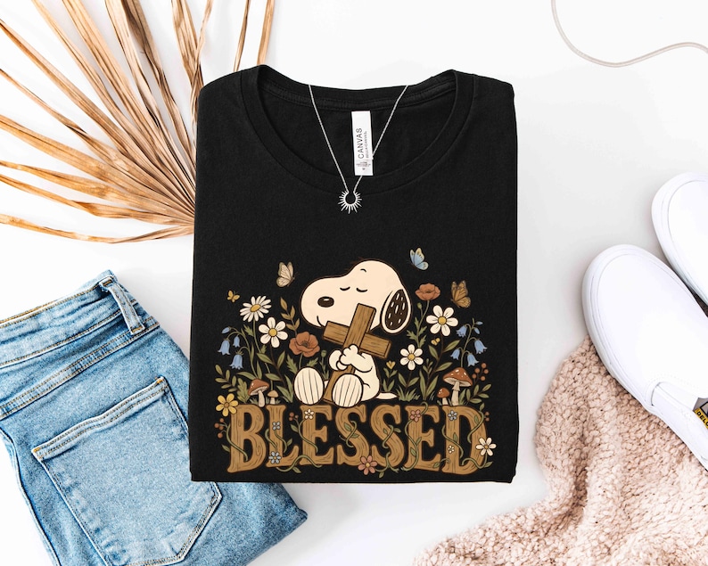 Puede incluir: Camiseta negra con Snoopy sosteniendo una cruz, rodeado de flores y mariposas. La palabra "BLESSED" se muestra en una fuente decorativa. Se ve un collar plateado con un colgante de sol.