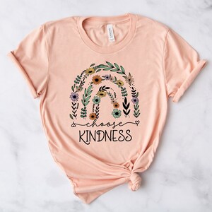 K&ouml;nnte beinhalten: Peachfarbenes T-Shirt mit einem floralen Regenbogen-Design und dem Text "choose kindness" in Schwarz.