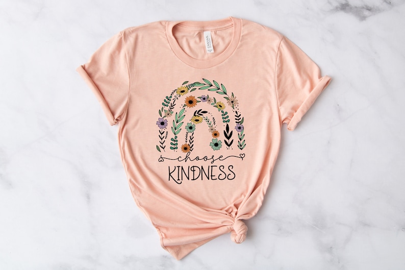 K&ouml;nnte beinhalten: Peachfarbenes T-Shirt mit einem floralen Regenbogen-Design und dem Text "choose kindness" in Schwarz.