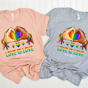 May include: Two t-shirts with a rainbow heart design and the text "Love is Love Pride". One shirt is peach and the other is gray.