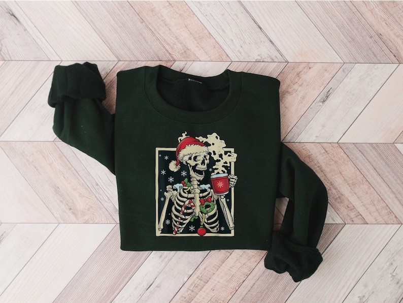 May include: A dark green sweatshirt with a festive skeleton graphic wearing a Santa hat and holding a mug of hot cocoa. The skeleton is surrounded by candy canes, holly, and snowflakes.